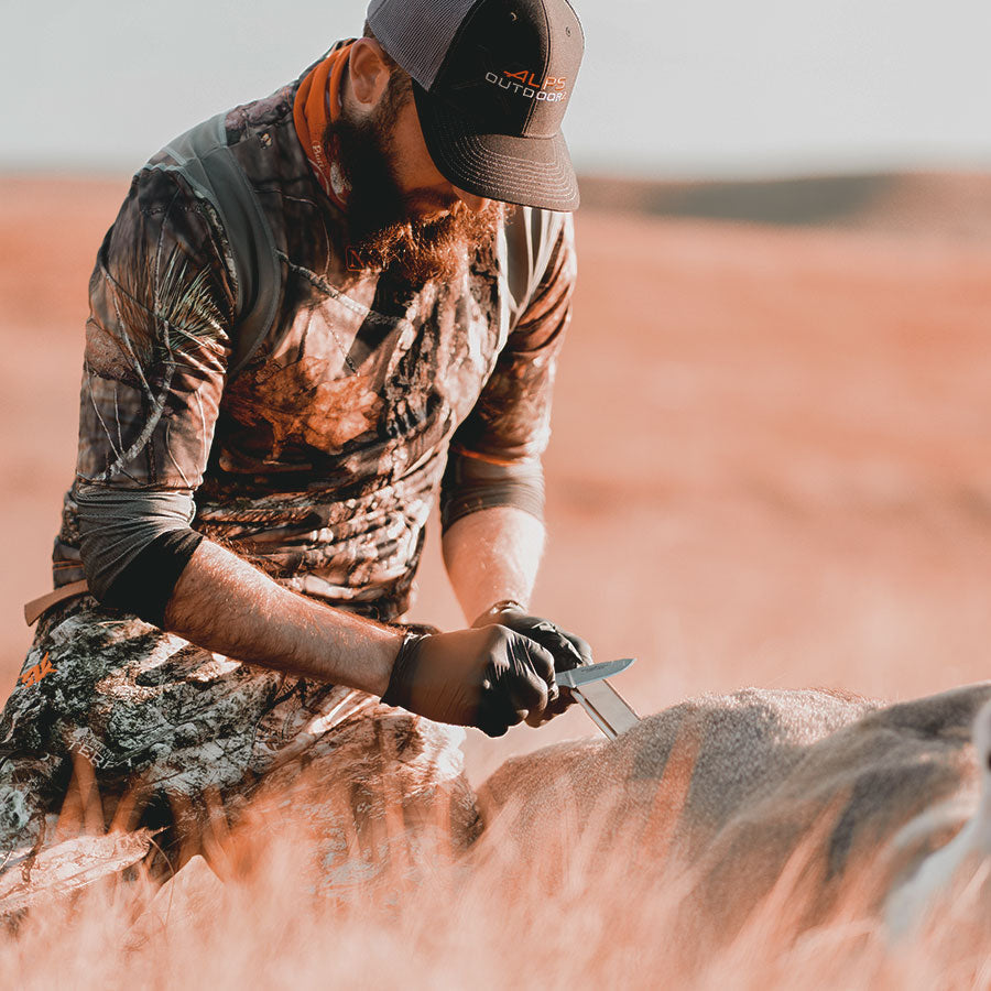 A man in camo leaning over a deer sharpening his hunting knife with a Work Sharp Guided Field Sharpener