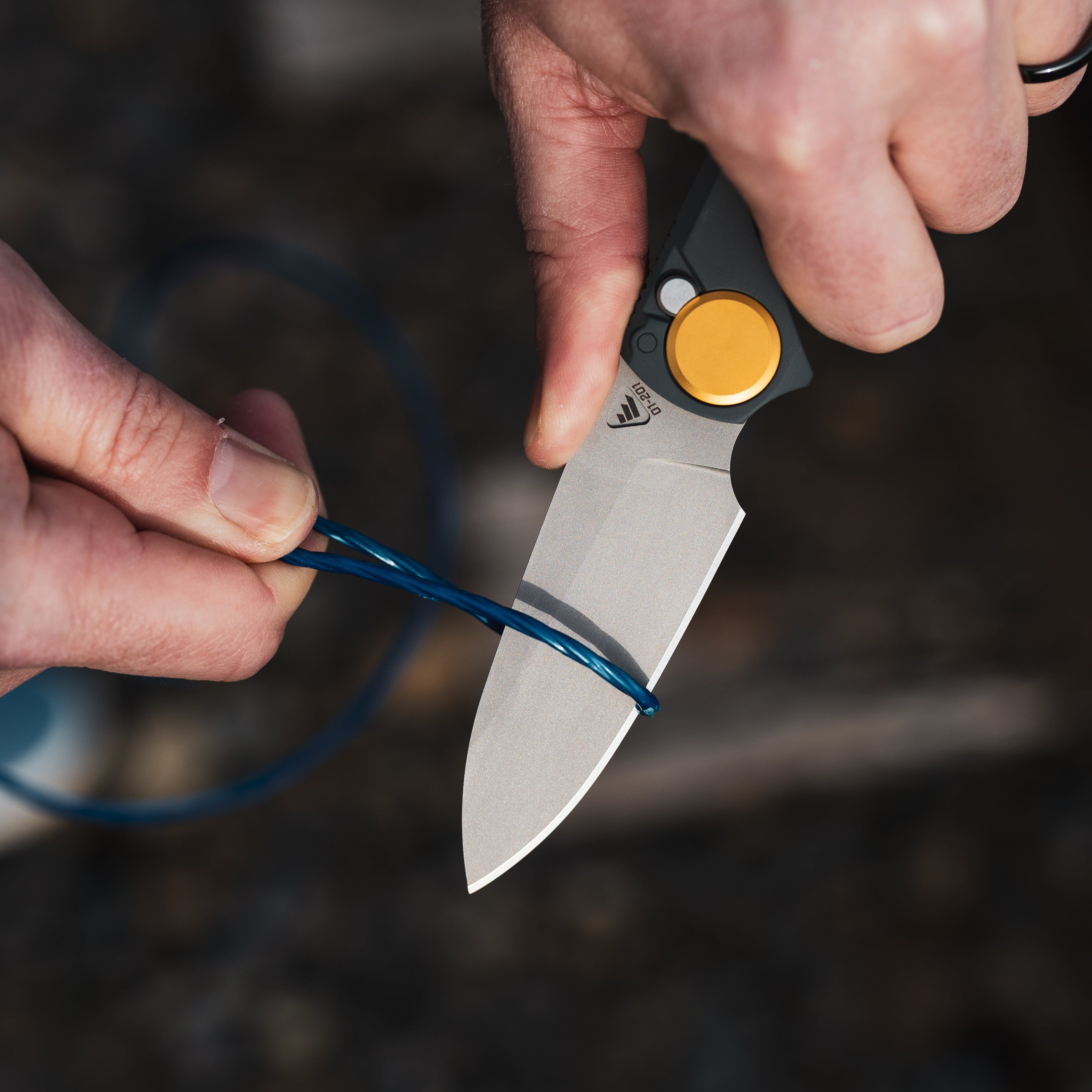 Person using a RMX Drop Point Compact knife to cut a blue cord with a blurred natural background
