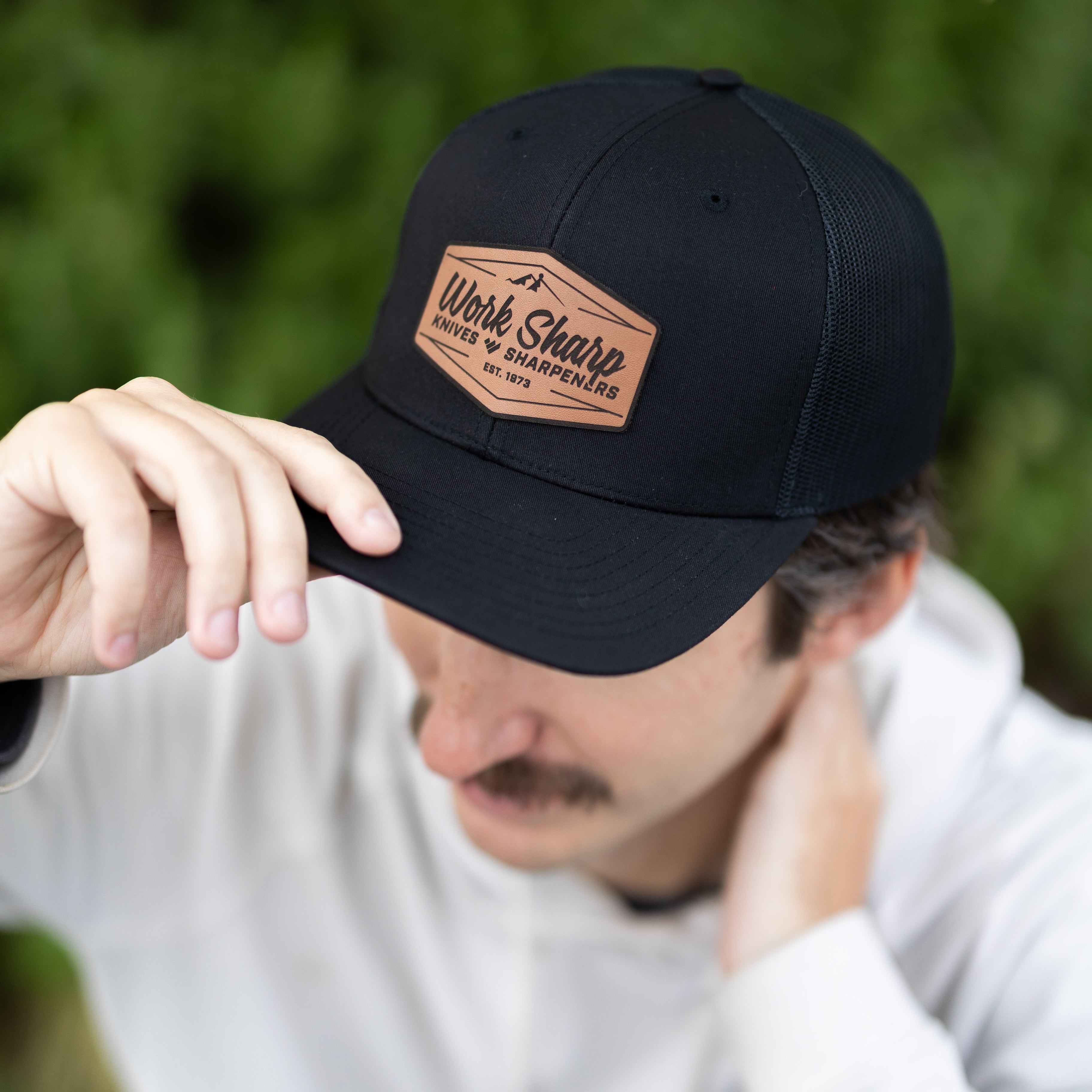 Person wearing a Work Sharp black cap with a leather patch, outdoors.