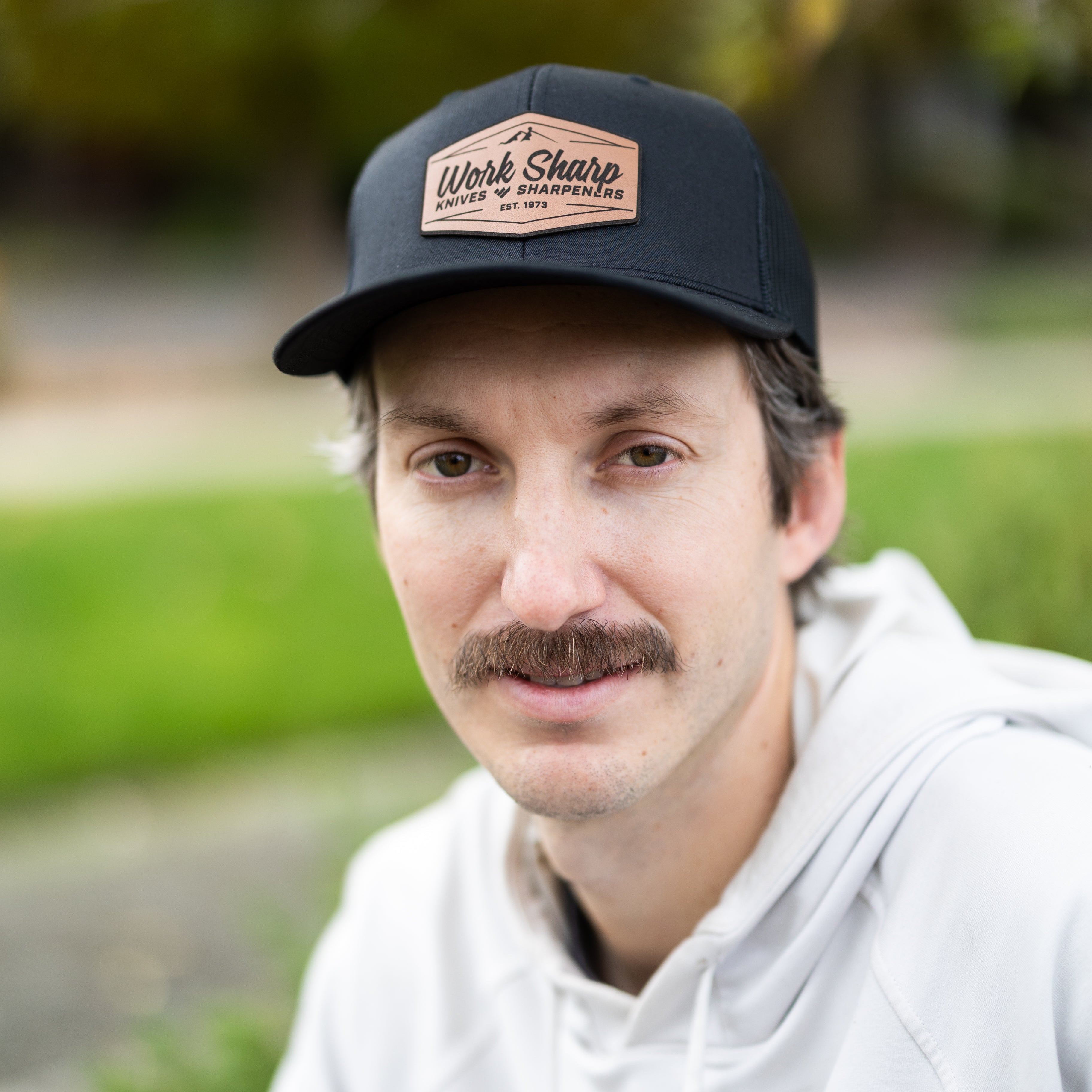 Man wearing a Work Sharp black cap with a logo, sitting outdoors with greenery in the background