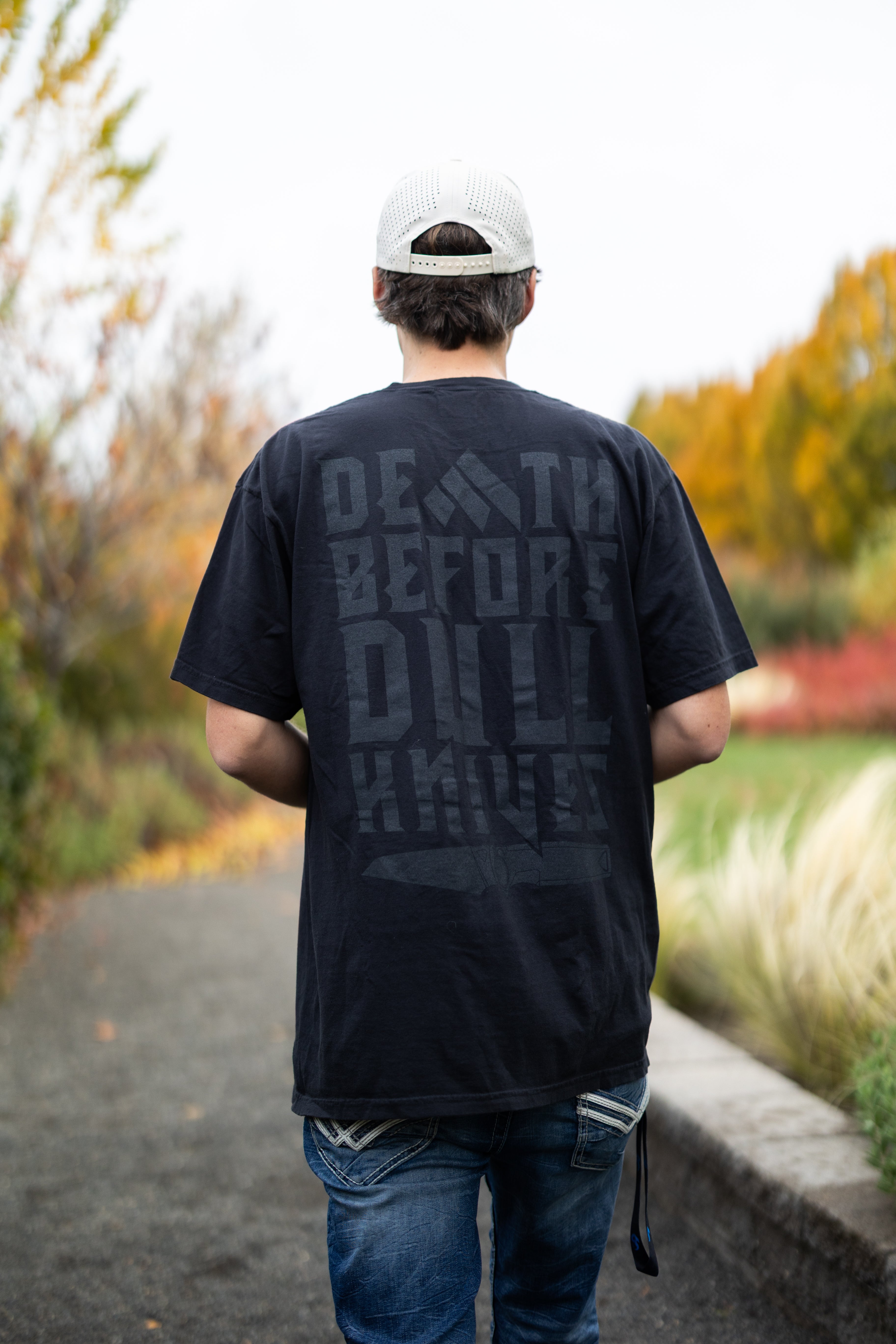 Person wearing a Work Sharp Death Before Dull Knives black t-shirt with text walking on a path in a park.