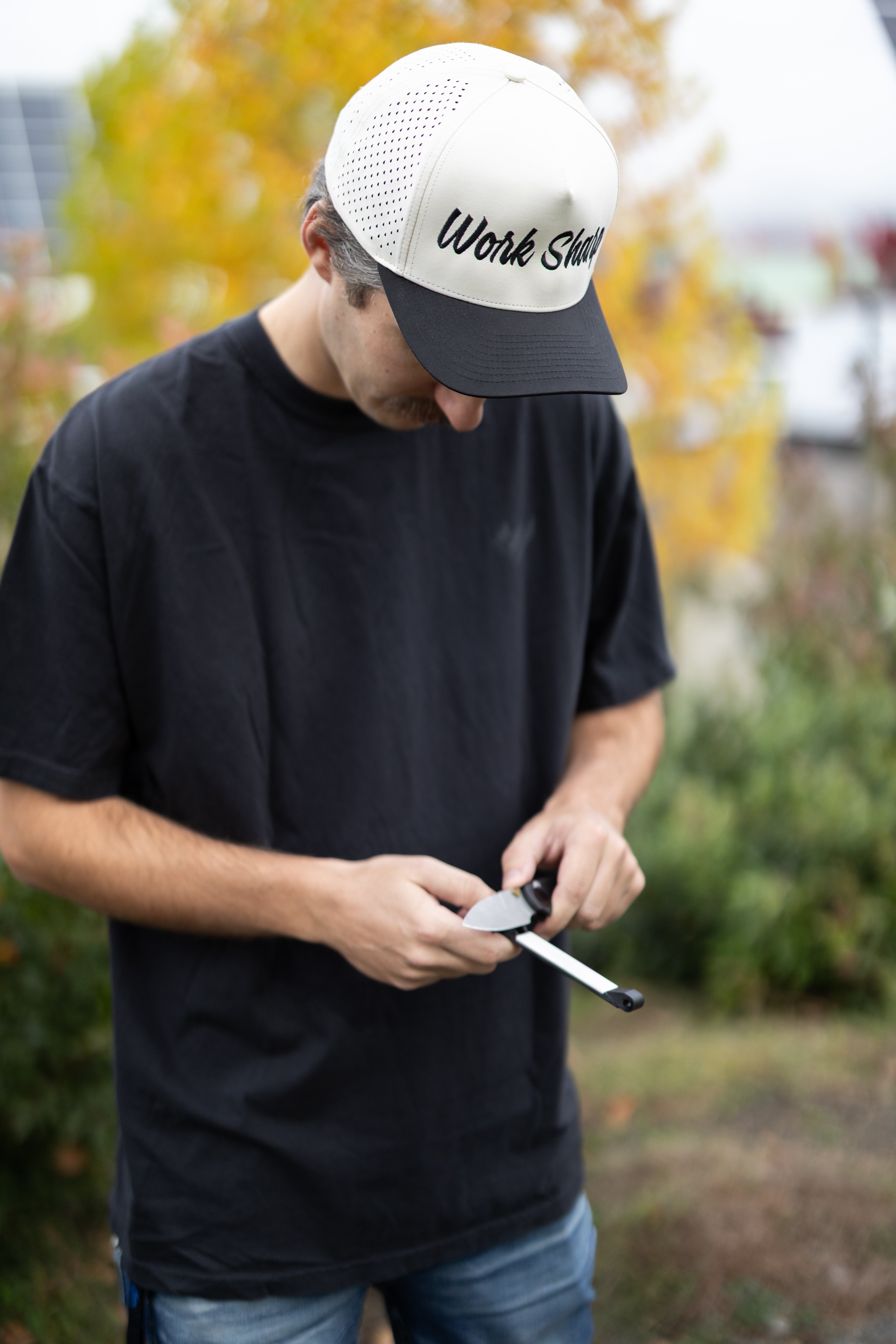 Man wearing a cap with 'Work Sharp Death Before Dull Knives' text, holding a tool outdoors.