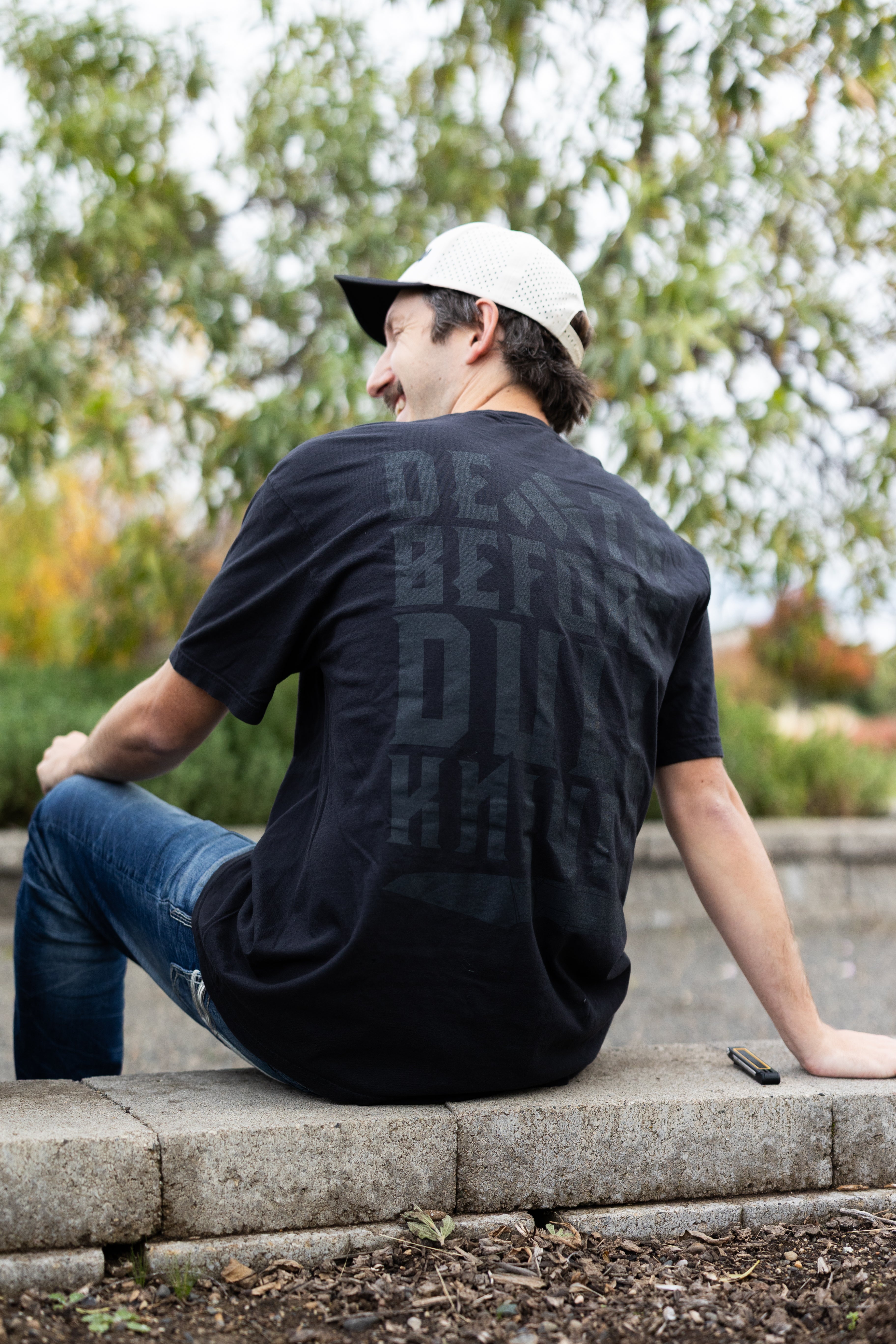 Person wearing a Work Sharp Death Before Dull Knives black t-shirt with text sitting on stone steps outdoors.