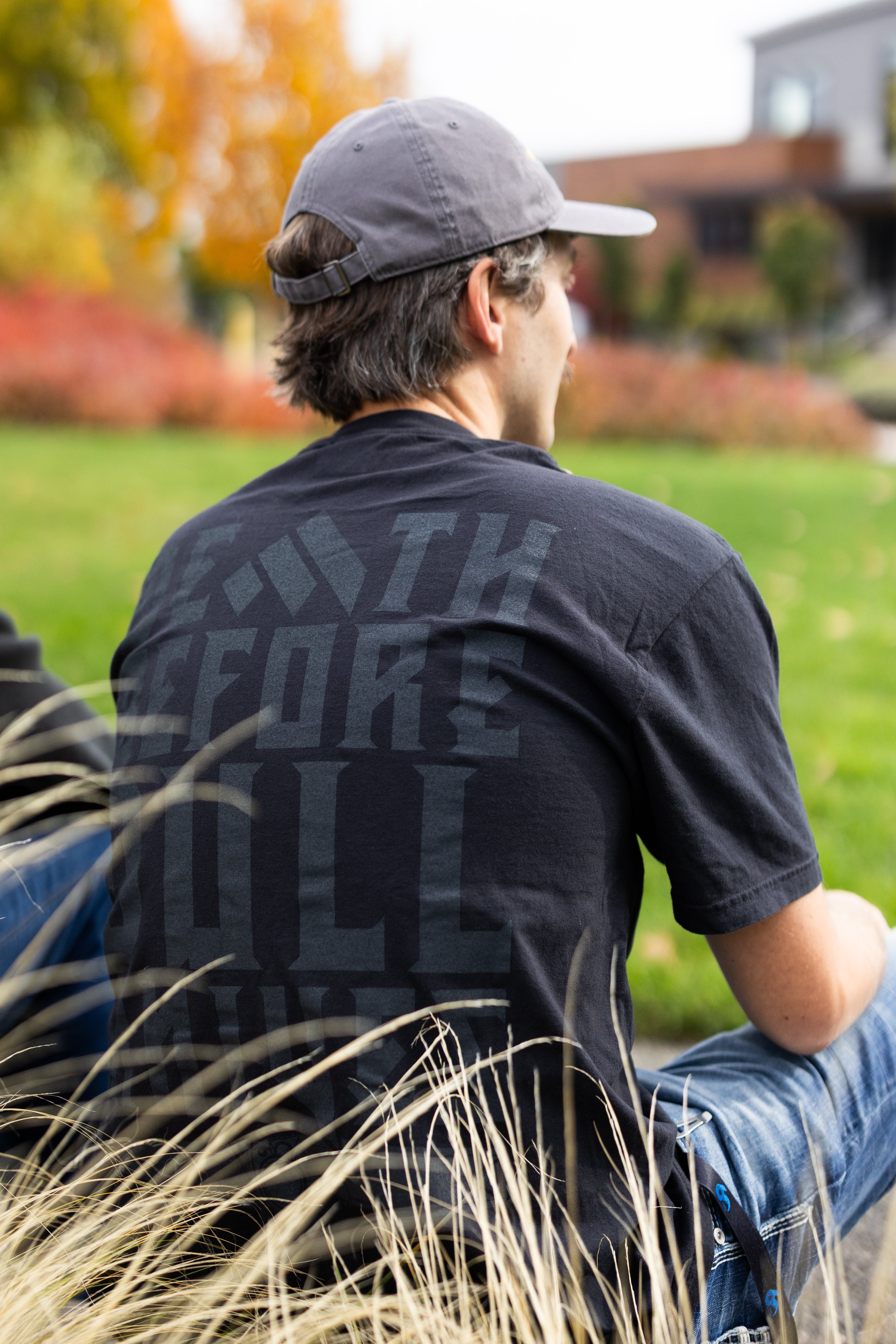 Person wearing a Work Sharp Death Before Dull Knives black t-shirt with text, sitting on grass with trees and buildings in the background