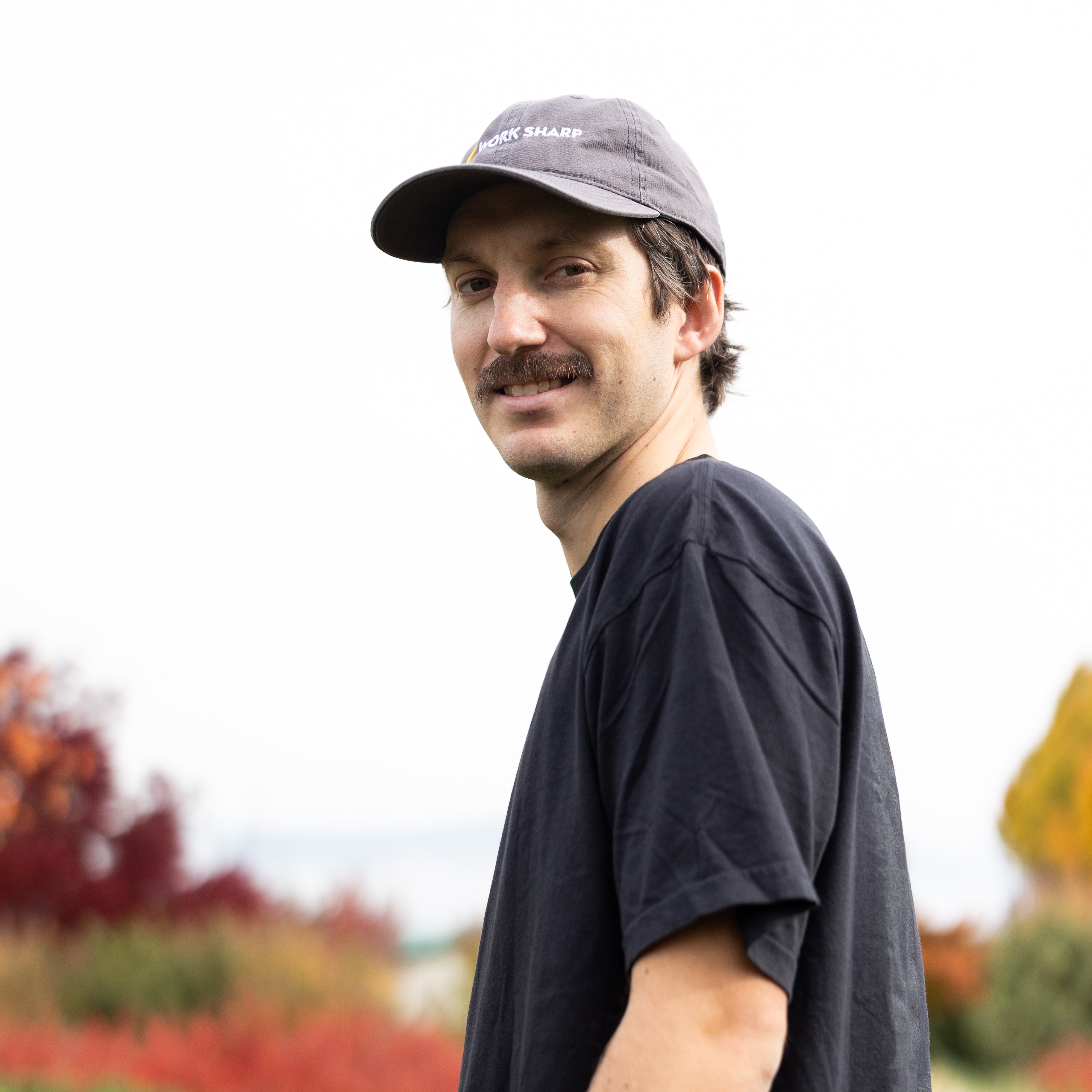Man wearing a black t-shirt and Work Sharp cap standing outdoors with trees in the background